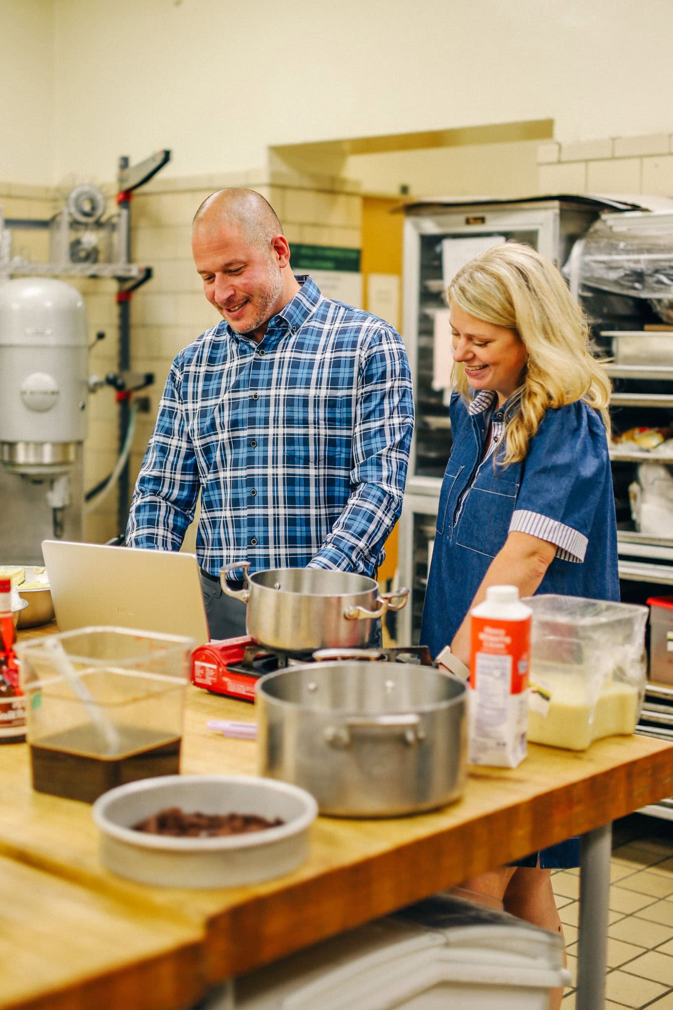 a man and a woman stand in a commercial kitchen and look at a laptop screen